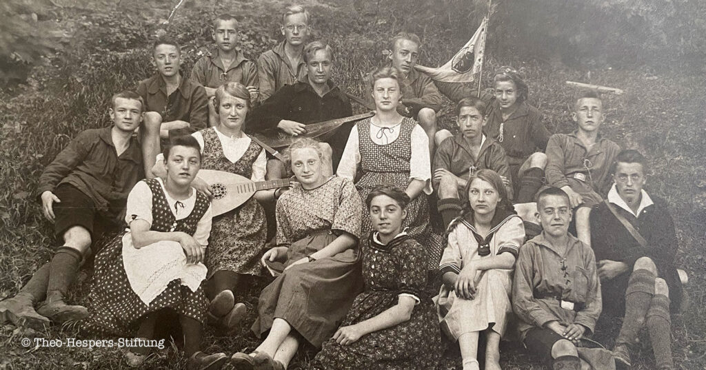 Ein Gruppenfoto von einer Quickborn-Gruppe in Mönchengladbach. In der Mitte thront beinahe Aenne Hespers, die ältere Schwester von Theo Hespers, der hinter ihr keck in die Kamera schaut. Er trägt ein Barrett und hält die Ecke eines Wimpels so, dass das Wappen auf dem Banner der Gruppe besser zu erkennen ist.